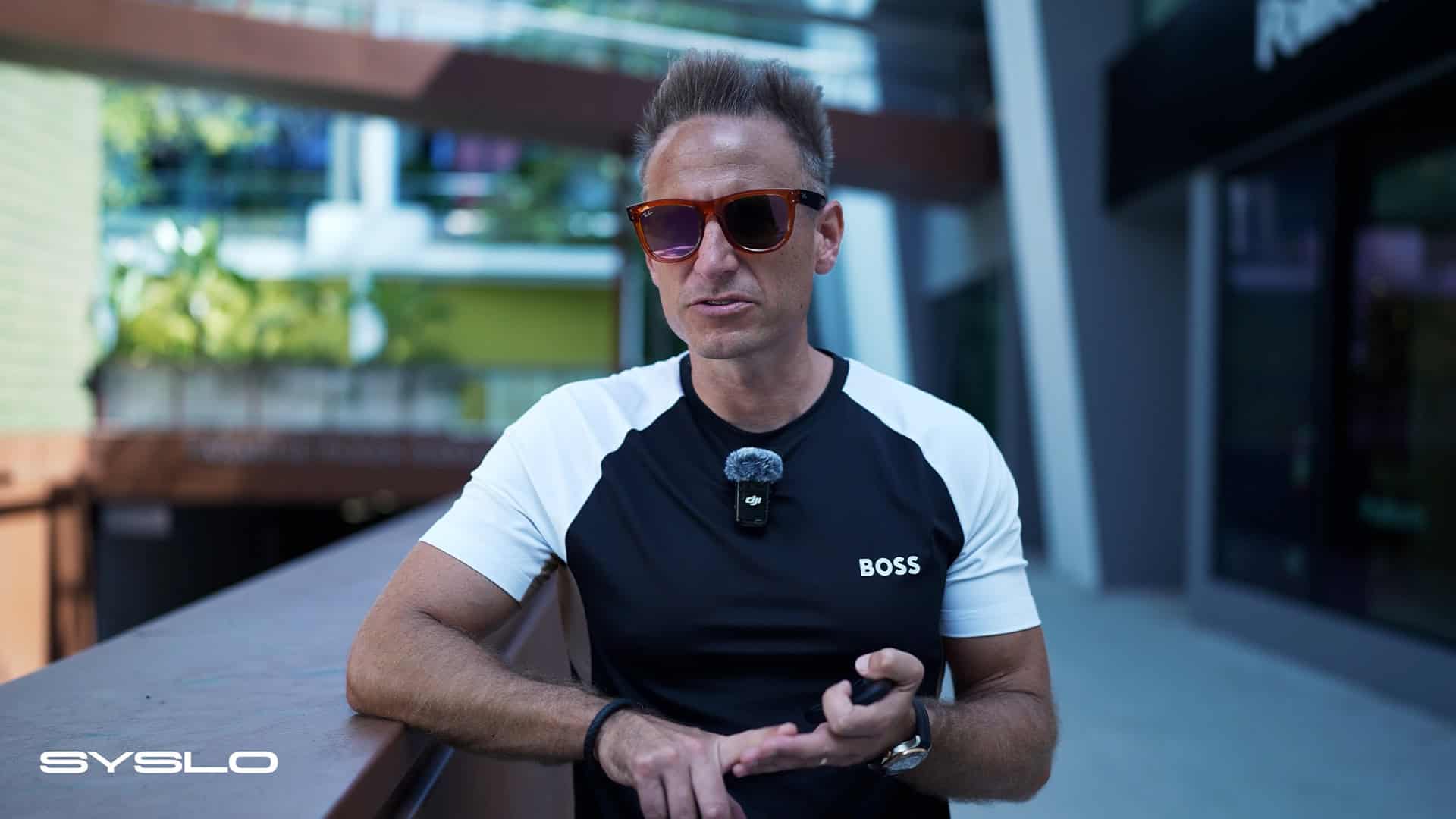 Professional businessman outdoors wearing sunglasses and a "BOSS" t-shirt, holding a smartphone, in a modern corporate environment representing innovation and business growth.