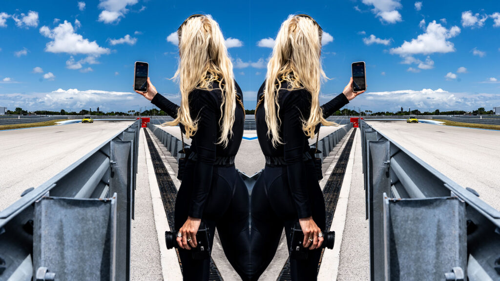 Female photographer at race track holding smartphone and camera, with edited mirrored effect, sunny day, blue sky with clouds, racing cars in background, professional motorsport photography, high-speed racing environment, dynamic action shot, motorsport event, trackside photography.