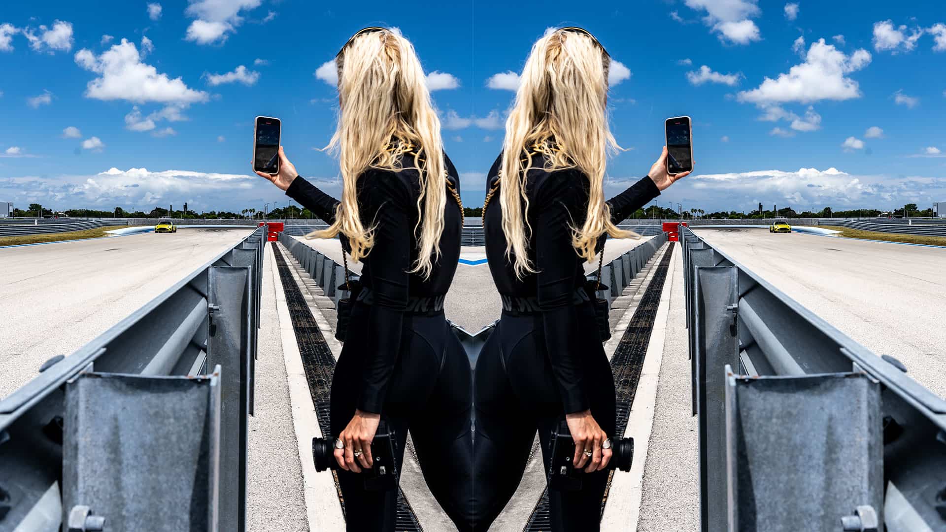 Female photographer at race track holding smartphone and camera, with edited mirrored effect, sunny day, blue sky with clouds, racing cars in background, professional motorsport photography, high-speed racing environment, dynamic action shot, motorsport event, trackside photography.