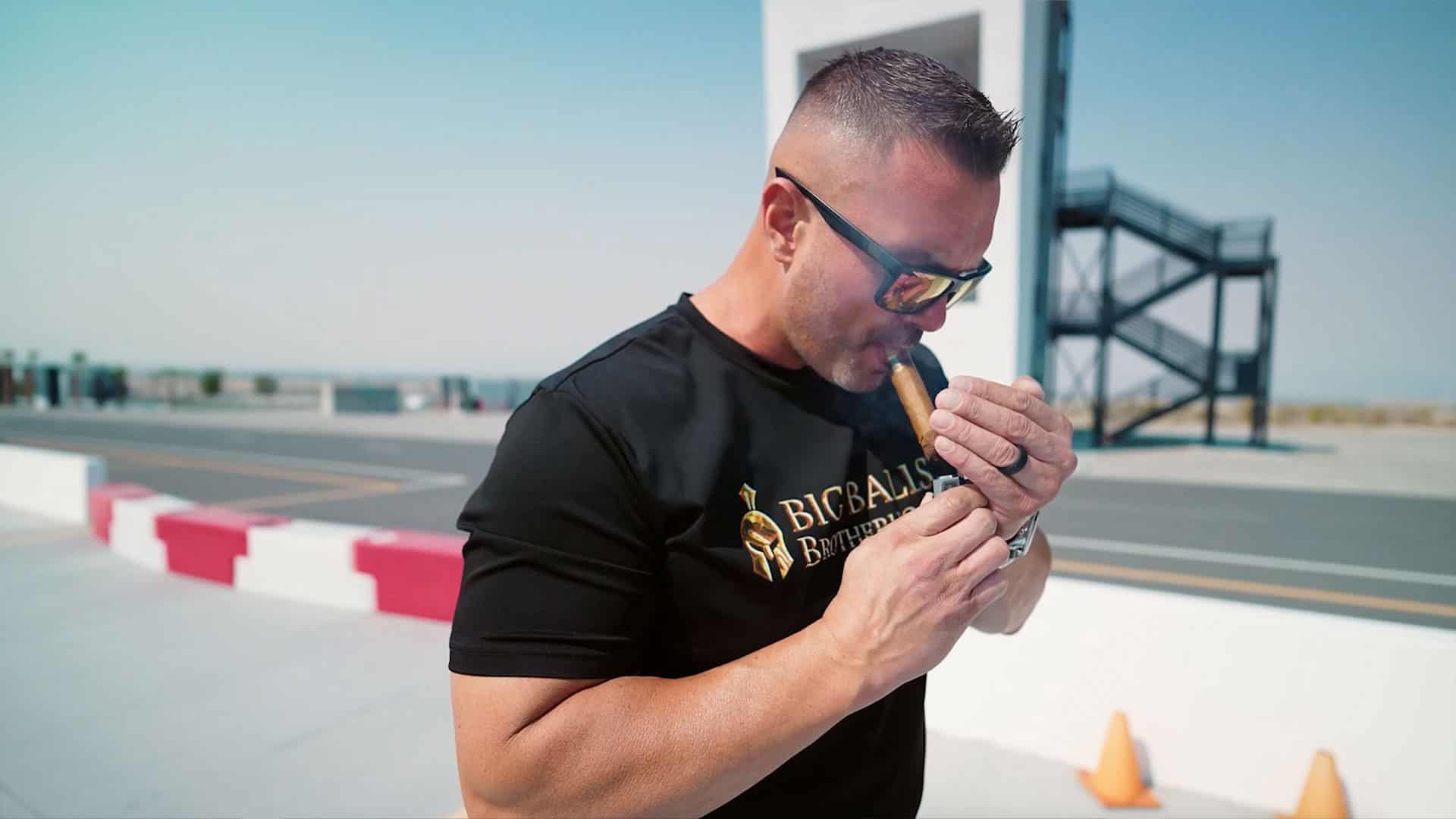 A man wearing sports sunglasses and a black T-shirt with "BICBALLS BROTHER" logo, smoking a cigar on a rooftop with a clear sky and a race track in the background.