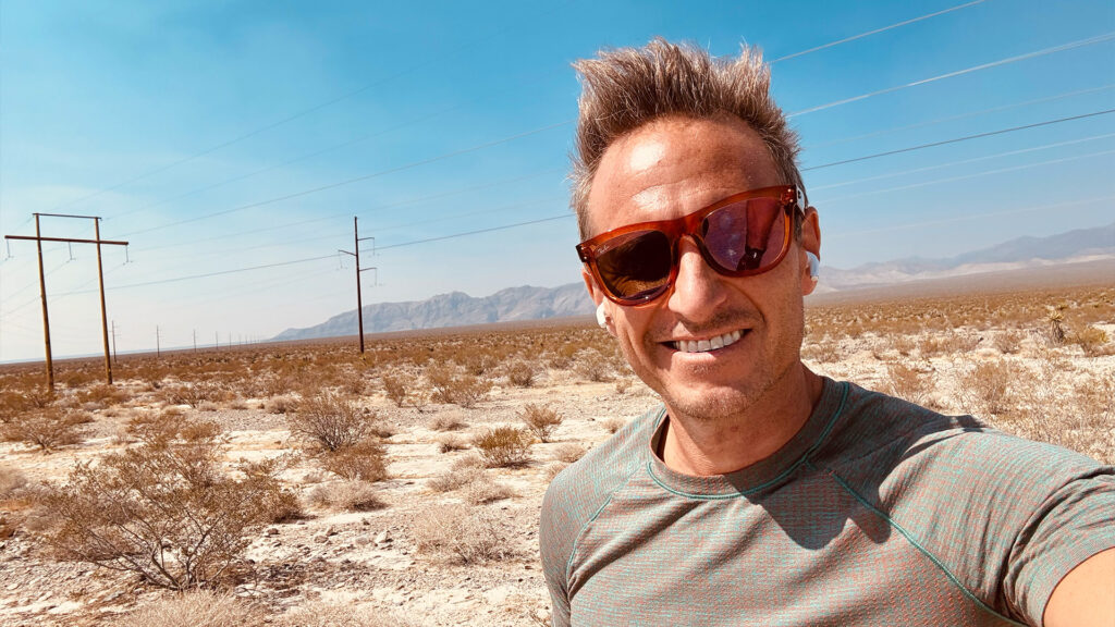 Vibrant selfie of a smiling man wearing sunglasses in a desert landscape with power lines and mountains, showcasing adventure and resilience, highlighting Syslo Ventures' focus on innovation and dynamic ventures.