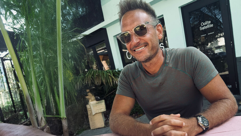 Vibrant smiling man sitting outdoors at a cafe in front of a modern building with lush greenery, showcasing luxury lifestyle and leisure, important for SEO in lifestyle and hospitality sectors.