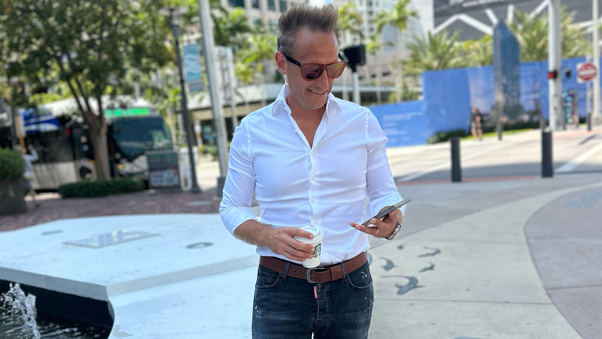 Modern businessman in white shirt with sunglasses holding coffee and smartphone in urban cityscape.