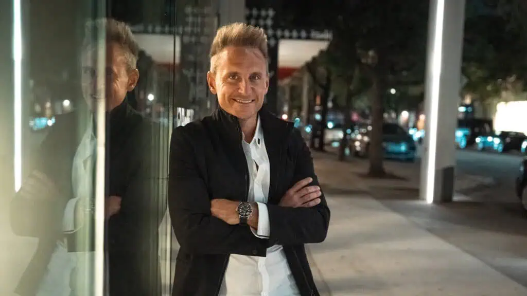 Bright smiling man standing confidently outside during night, wearing a white shirt and black jacket, with a city street background featuring parked cars and illuminated trees, representing entrepreneurial success and modern business ambiance.