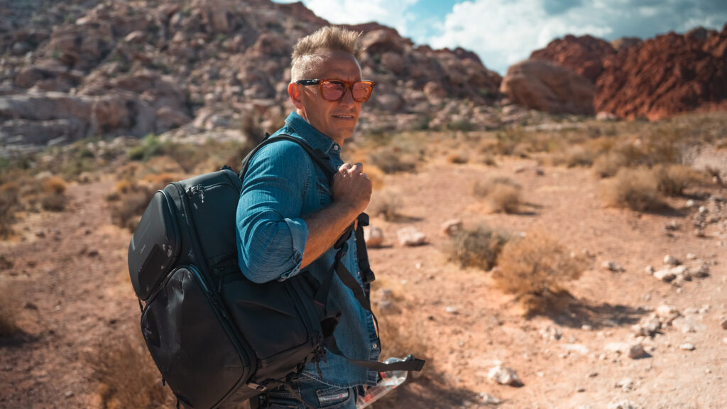 Traveling man with backpack hiking through desert landscape with red rocks and blue sky, wearing sunglasses, exploring outdoor adventure, representing outdoor gear and adventure travel.