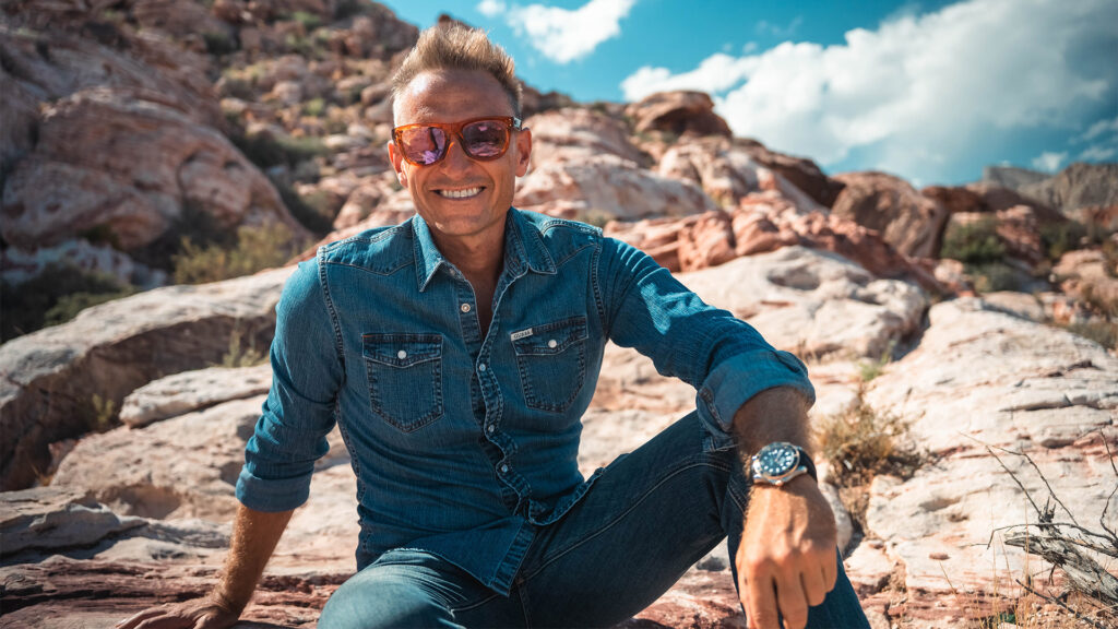Vibrant outdoor portrait of a smiling man in sunglasses and denim shirt sitting on rocky terrain in a desert landscape, showcasing adventure, confidence, and outdoor lifestyle.
