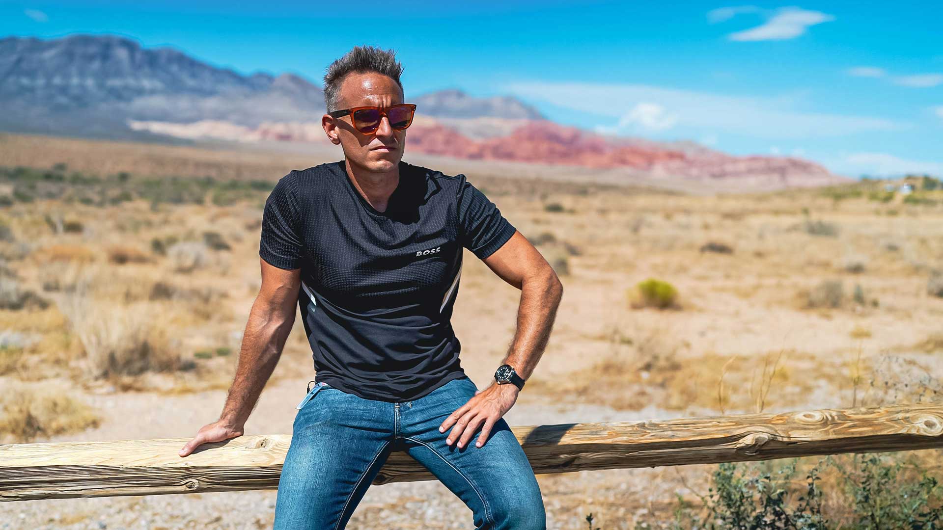 Vibrant desert landscape with mountains in the background, featuring a stylish man in casual wear leaning on a wooden fence, highlighting adventure, outdoor lifestyle, and exploration themes.