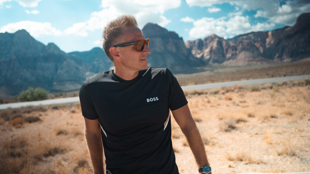 Professional man wearing sunglasses and a "BOSS" T-shirt outdoors in a desert mountain landscape, representing adventure and corporate style.