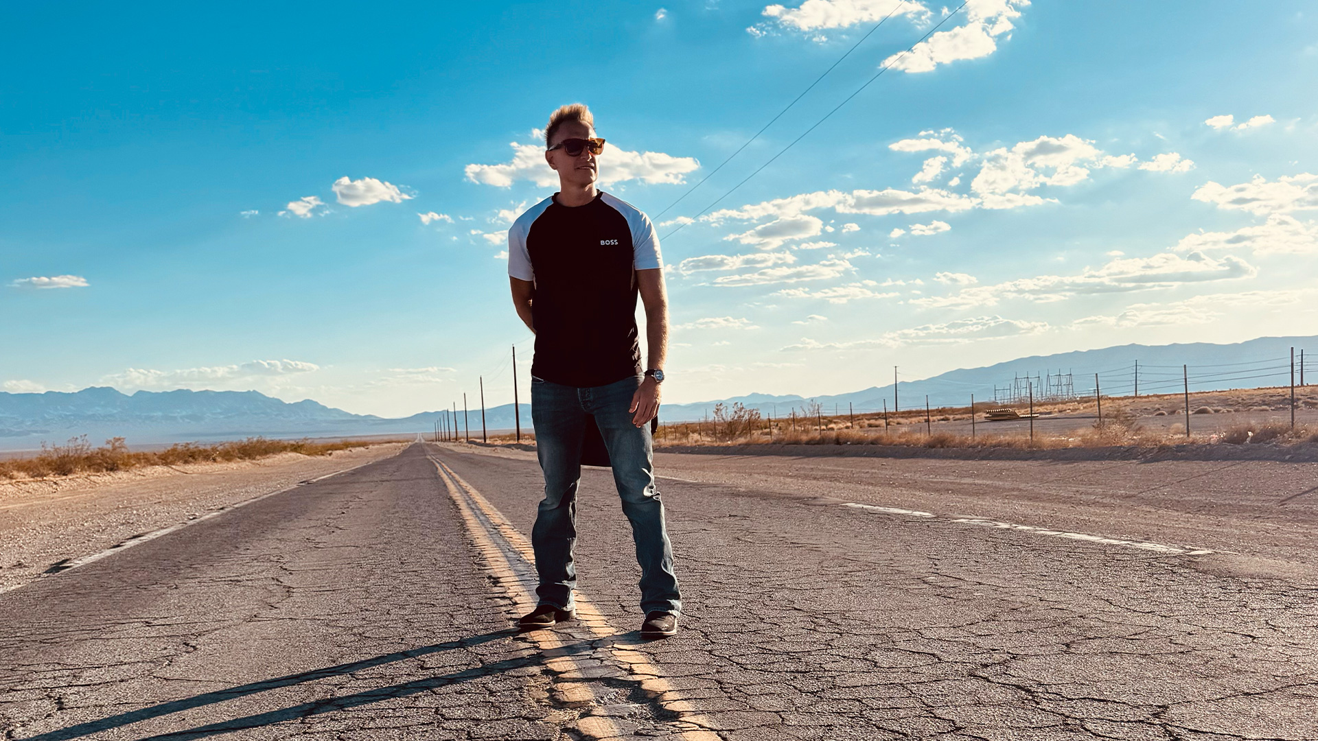 Empty desert highway with a man in casual attire standing in the middle, mountains in the background, clear blue sky, and power lines, representing adventure, exploration, and freedom.