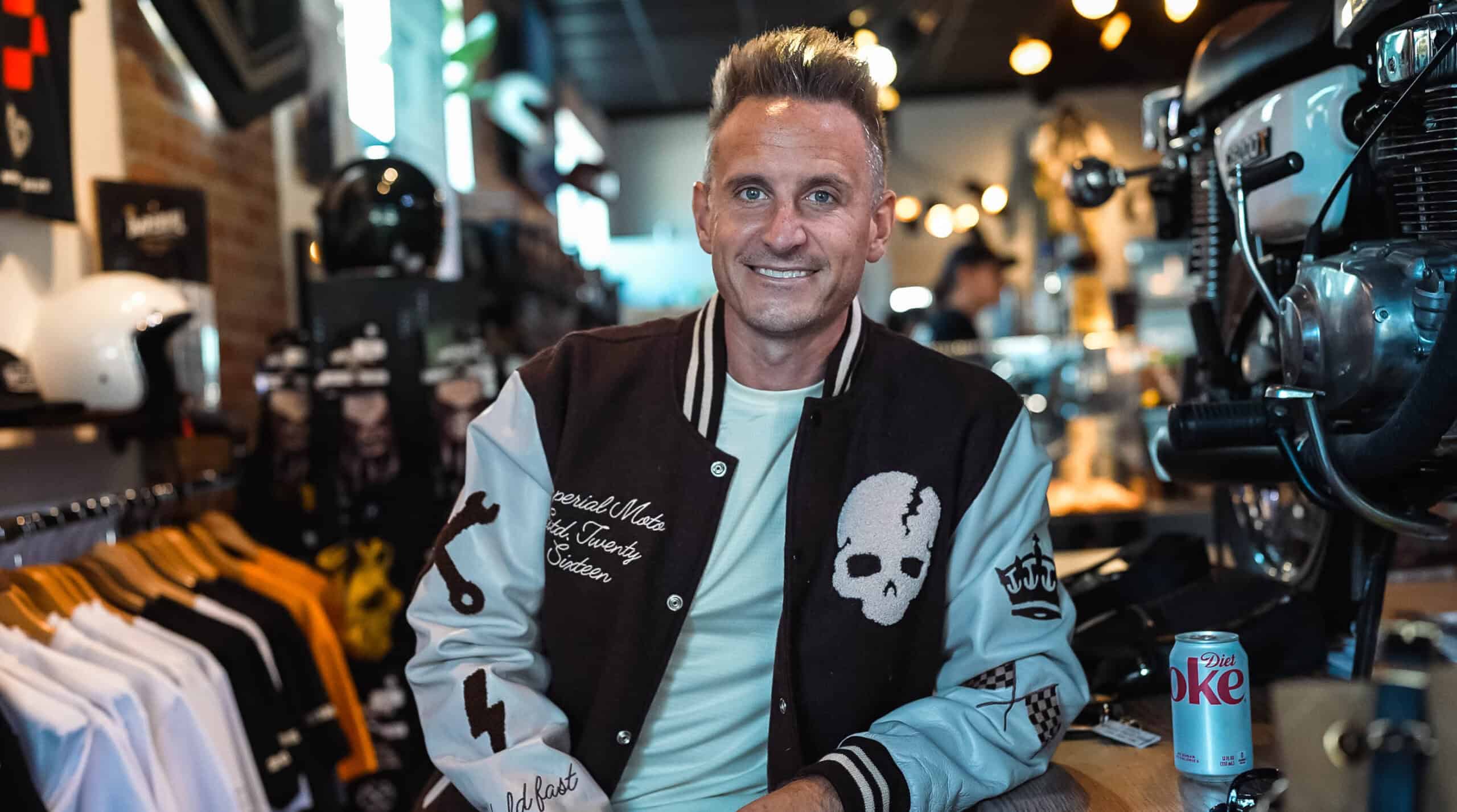Stylish man smiling inside motorcycle apparel shop, wearing trendy varsity jacket, surrounded by motorcycle helmets, gear, and apparel, representing motorcycle lifestyle and fashion.