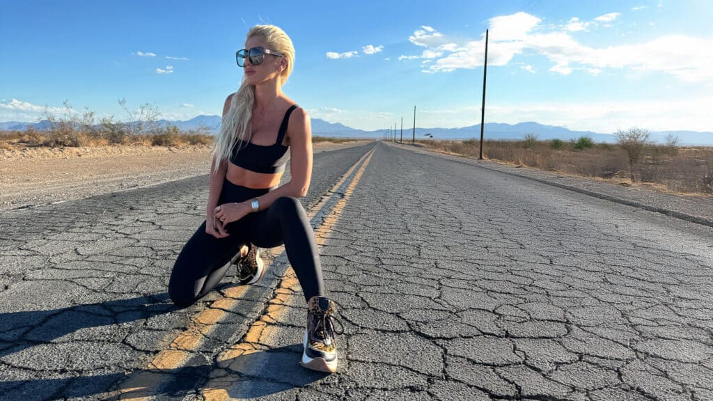 Kneeling woman in athletic wear on an empty desert road with mountains in the background, symbolizing adventure, fitness, and resilience for innovative business ventures.