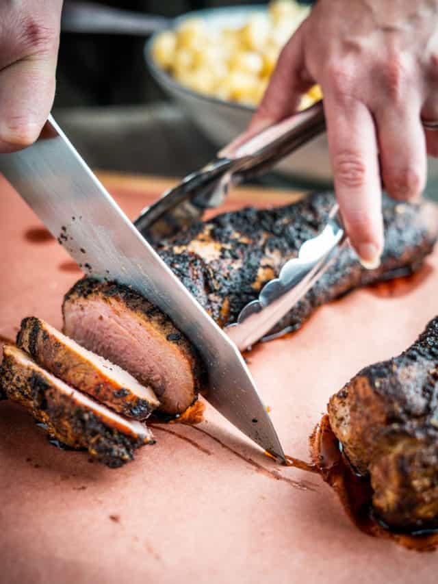 Chef slicing a cooked beef tenderloin with a sharp knife on a wooden cutting board.
