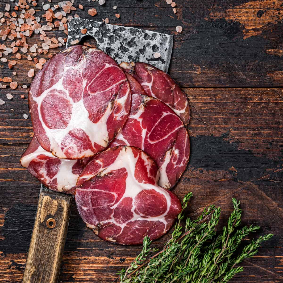 thinly sliced Capicola on a wood background with fresh herbs and pink sea salt