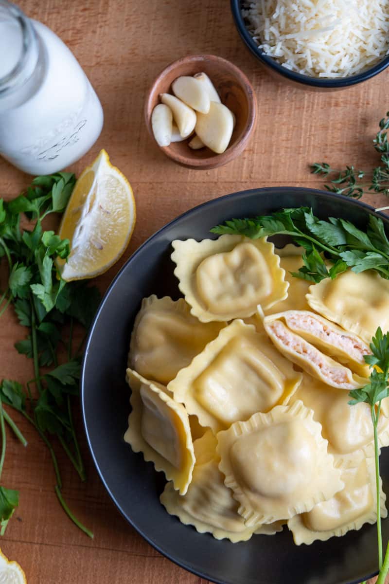 lobster ravioli, one cut so you can see the pink inside on a table with garlic, lemon wedges and cream