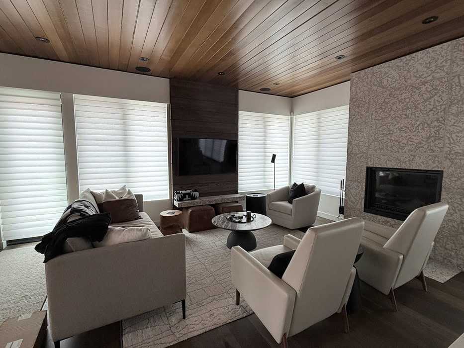 Bright modern living room with large windows and white blinds in Clear Lake, MB. Comfortable seating area with sofas, armchairs, and a fireplace creates a warm, inviting atmosphere.