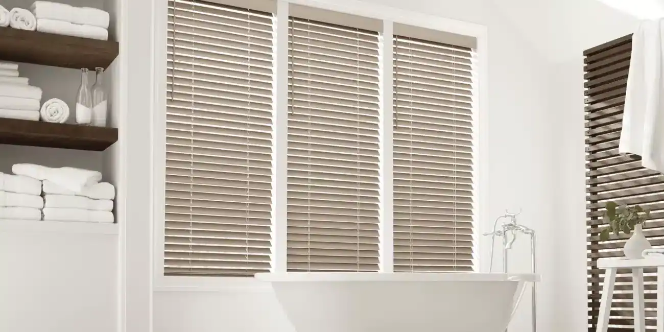 Bright bathroom featuring wooden horizontal blinds on a large window, with white walls, towels, and minimalistic decor for a clean and relaxing atmosphere.