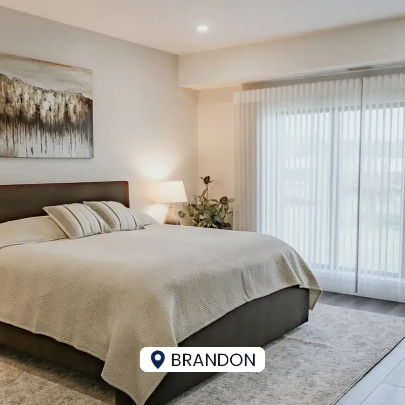 A cozy bedroom featuring a large window with sheer curtains, a comfortable bed with neutral bedding, and modern wall art, creating a peaceful and inviting atmosphere.