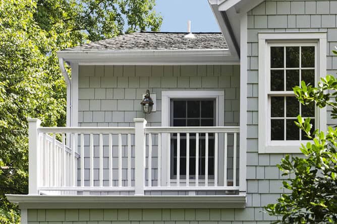 A lush green tree and a white balcony railing on a light grey house exterior.