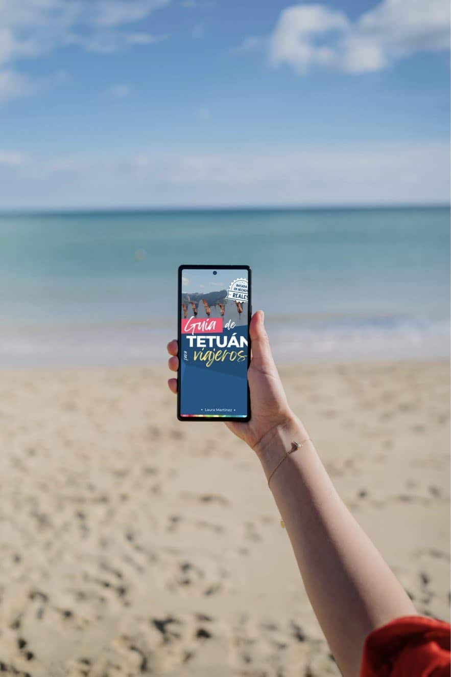 Persona sosteniendo un teléfono en la playa con guía de Tetuán en pantalla.