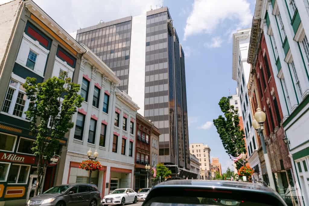 A vibrant street scene in downtown Roanoke, Virginia, showcasing a mix of historic and modern architecture. Colorful storefronts with flower baskets line the street, while a tall, glass-fronted building towers above, contrasting with the older, low-rise buildings. The bright sky and bustling street capture the blend of old and new in Roanoke's cityscape.