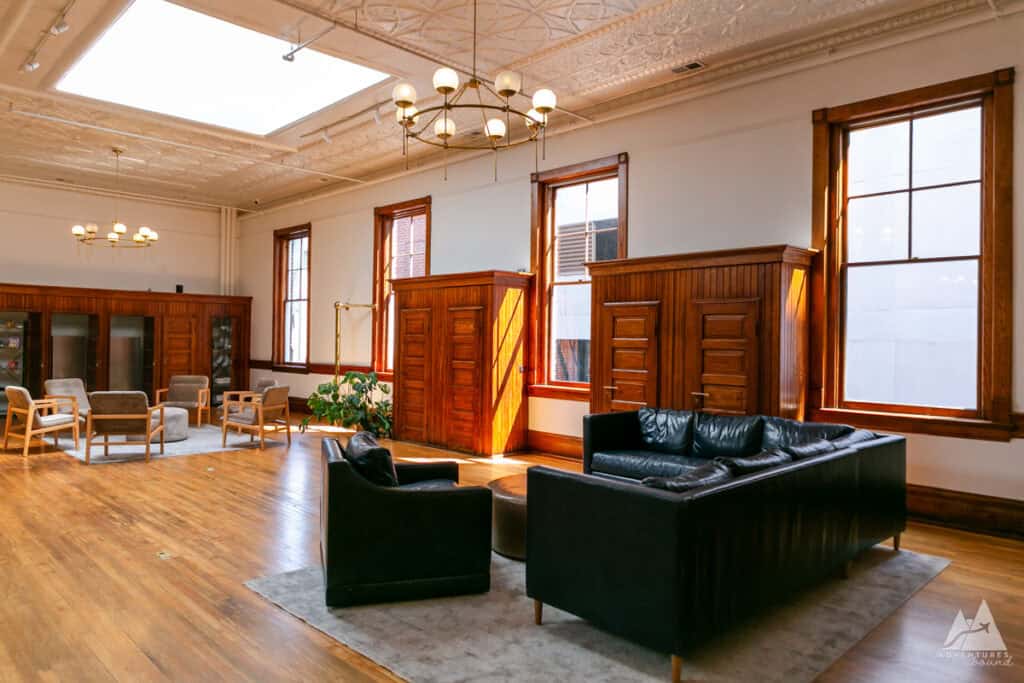 A spacious lounge area in the Fire Station One Boutique Hotel, featuring high ceilings with intricate molding, large windows, and polished wood floors. The room is furnished with black leather couches, wooden cabinets, and modern armchairs arranged for casual seating, illuminated by warm natural light from skylights. The blend of vintage architecture and contemporary furnishings creates a stylish, comfortable gathering space.