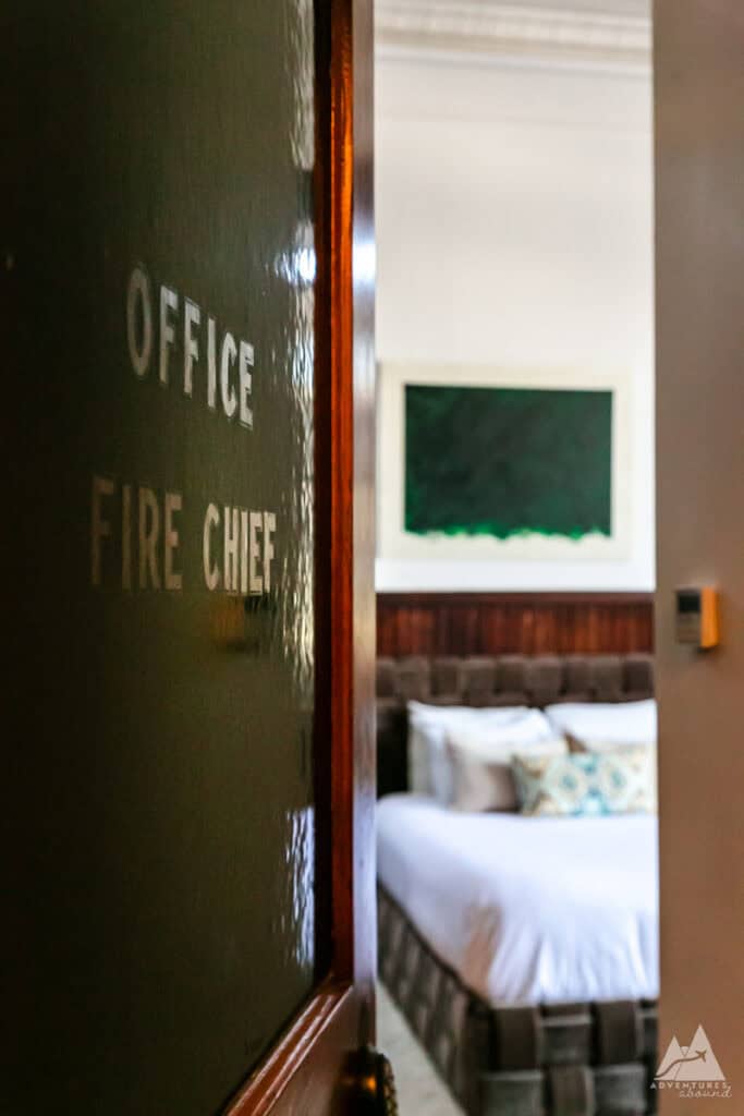 A partially open door labeled "Office Fire Chief" reveals a glimpse into a boutique hotel room at Fire Station One. Inside, a plush bed with patterned pillows and a deep green artwork on the wall create a stylish yet historic ambiance, hinting at the building’s firehouse origins. The scene combines modern comfort with vintage charm.