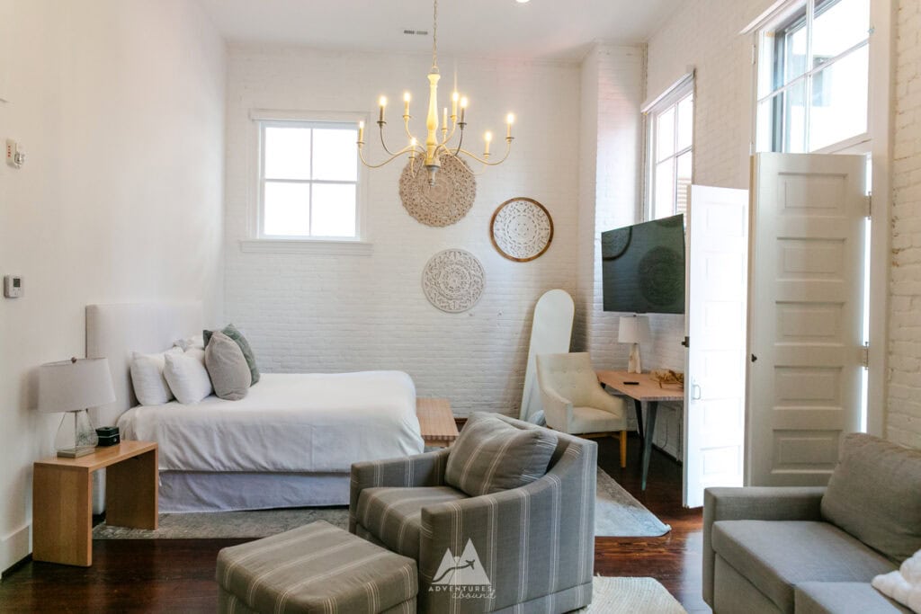 A bright, airy hotel room with white brick walls, a cozy bed with plush pillows, and a chandelier overhead in the Fire Station One Boutique Hotel. The room features an elegant mix of furniture, including a striped armchair, a desk with a modern chair, and woven wall decor, creating a relaxing and stylish atmosphere. Large windows let in natural light, enhancing the minimalist, cozy aesthetic of the space.