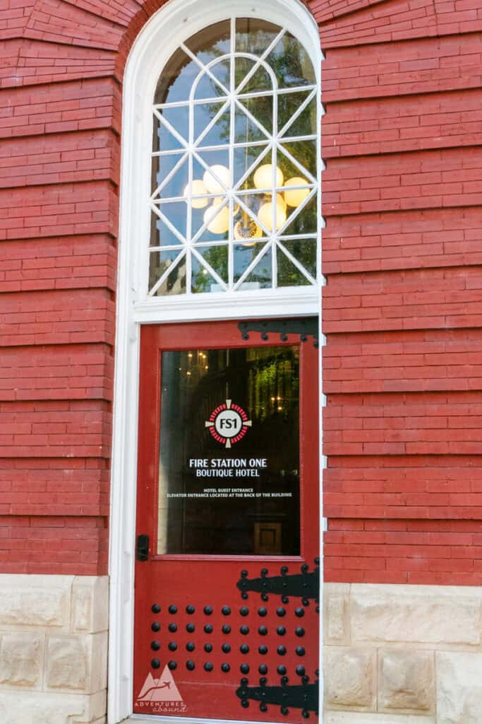 A close-up of the entrance to the Fire Station One Boutique Hotel in Roanoke, Virginia, featuring a red door with black rivet details and a tall arched window above. The door bears the hotel's logo and name, “Fire Station One Boutique Hotel,” along with directions for guests. The historic red brick exterior and classic design highlight the building’s original firehouse architecture, now repurposed as a boutique hotel.