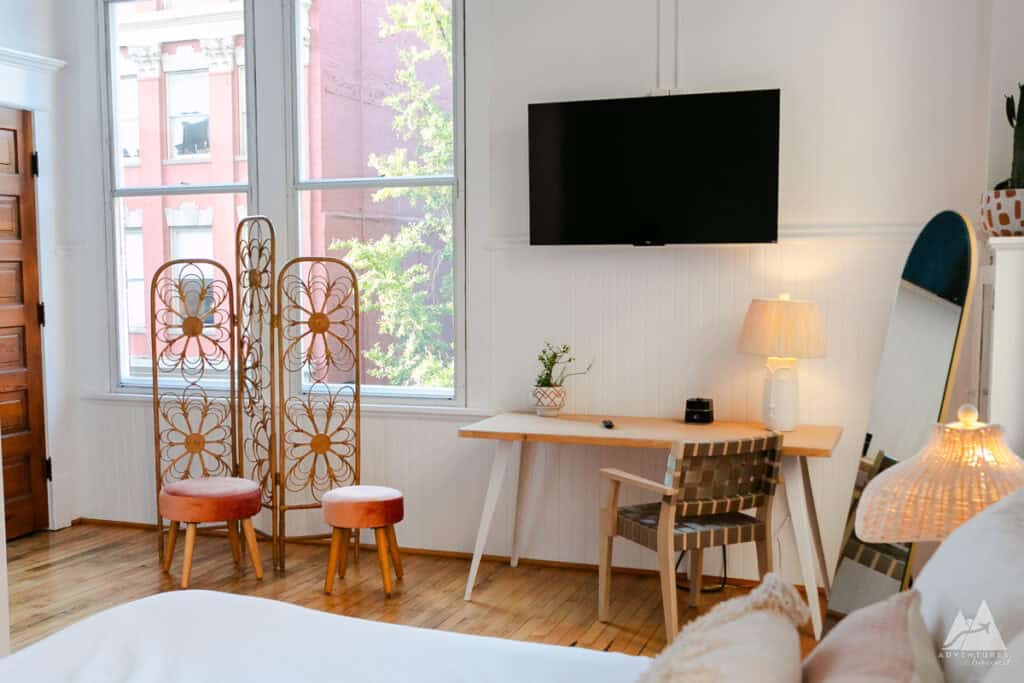 A stylish corner of a hotel room at Fire Station One Boutique Hotel, featuring large windows that let in natural light, a TV mounted on the wall, and a wooden desk with a woven chair. Decorative accents include a unique floral room divider with orange upholstered stools, a floor mirror, and a table lamp that adds warmth to the space.