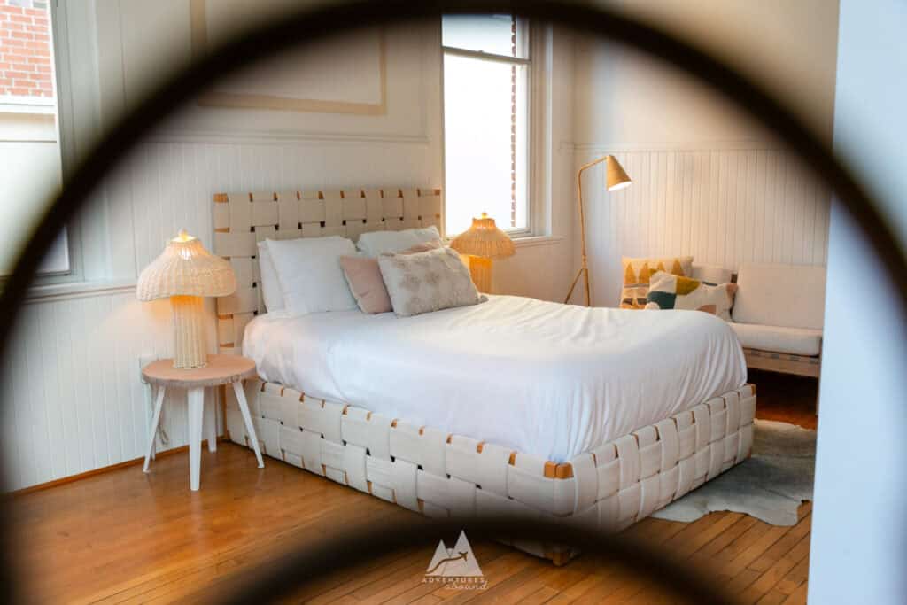 A cozy, minimalist bedroom in the Fire Station One Boutique Hotel, featuring a woven beige bed frame with crisp white linens, flanked by two wicker bedside lamps. A stylish floor lamp and decorative pillows add warmth to the space, which has white paneled walls and natural wood floors. The photo captures the room through a blurred foreground frame, creating a soft, inviting ambiance.