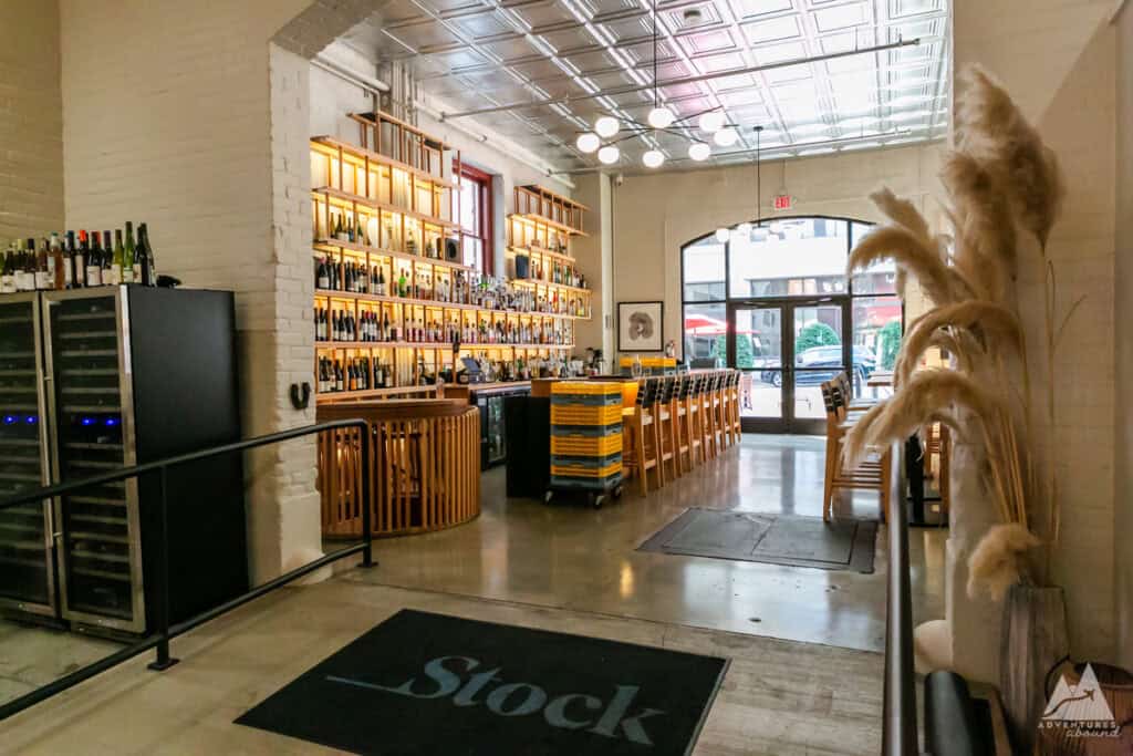 The interior of Stock Bistro restaurant in Roanoke, Virginia, featuring a sleek bar lined with wine bottles on backlit wooden shelves and a row of high chairs facing the counter. Modern lighting hangs from a reflective ceiling, and a cozy seating area with tall pampas grass adds warmth to the industrial-style decor. The glass doors at the far end provide a glimpse of the outdoor patio area, creating a bright and inviting dining atmosphere.