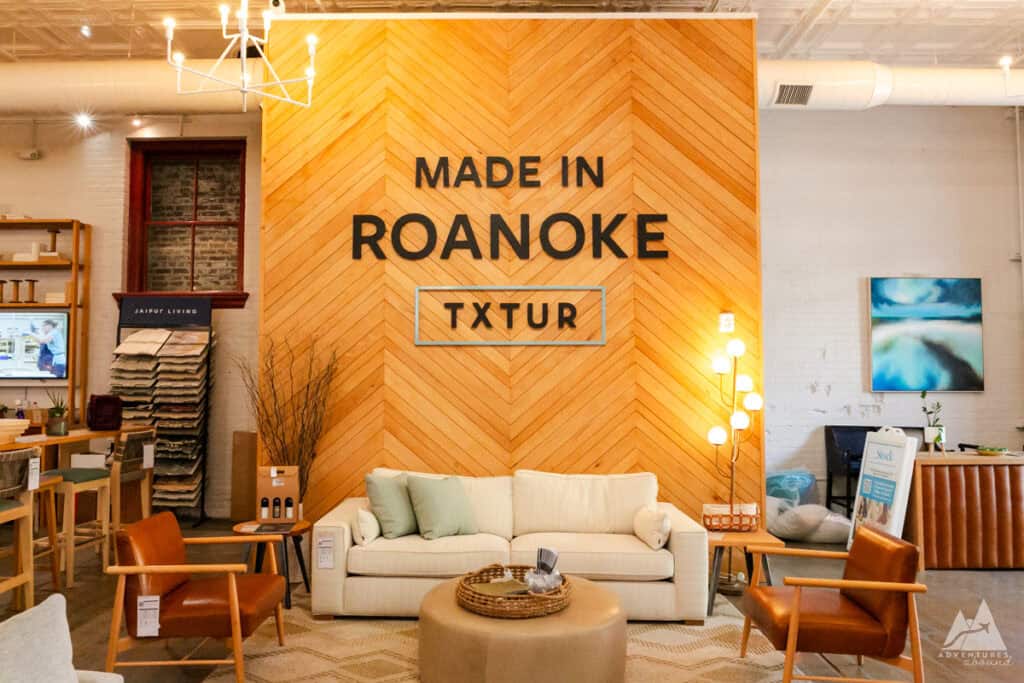 A stylish interior display with a large wooden chevron-patterned wall featuring bold text that reads "Made in Roanoke TXTUR," highlighting local craftsmanship. The space includes modern furniture like a cream sofa, leather chairs, and a central woven basket, creating a cozy, inviting showroom ambiance. The setup reflects a blend of rustic and contemporary design elements, emphasizing local production and design