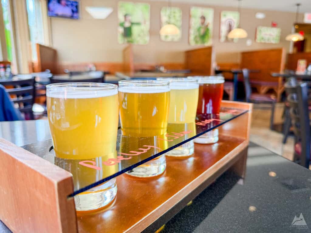 A wooden tray holds a flight of four beers, each in a small glass with varying shades from pale yellow to deep amber at Robinhood Brewing. The flight rests on a reflective black countertop, with a casual dining setting in the background, including booths, hanging pendant lights, and wall art.