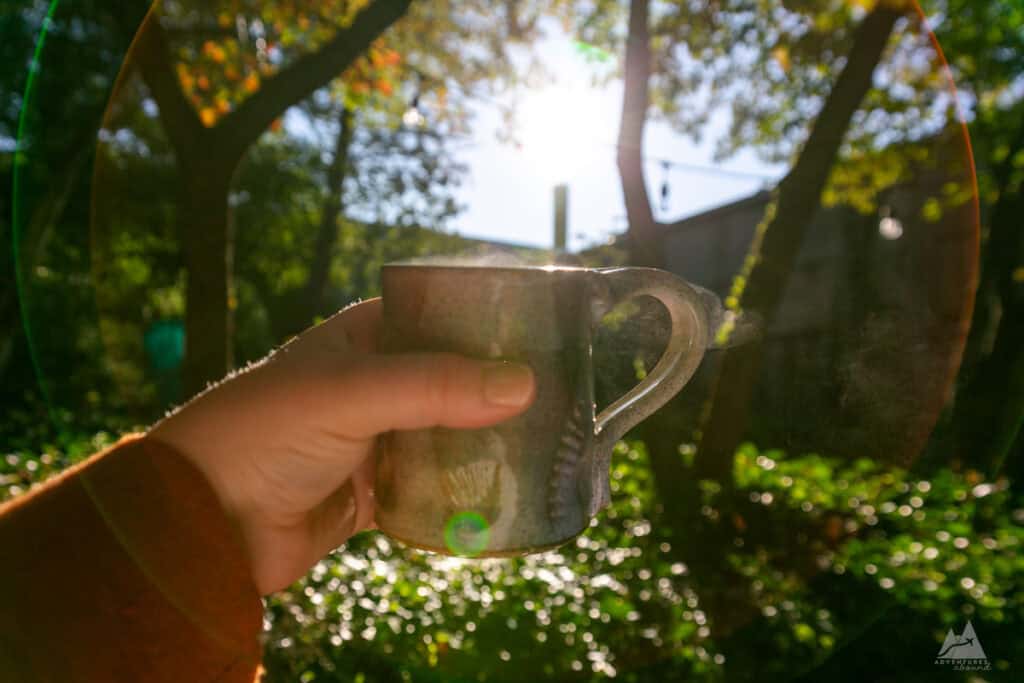 Morning coffee at Bellefonte Bed and Breakfast. A hand holds a ceramic mug with steam rising from its surface, catching the morning sunlight. The background features a lush green outdoor space with trees, string lights, and a soft sun flare effect. The warm, golden light enhances the cozy and peaceful atmosphere of enjoying coffee outdoors.