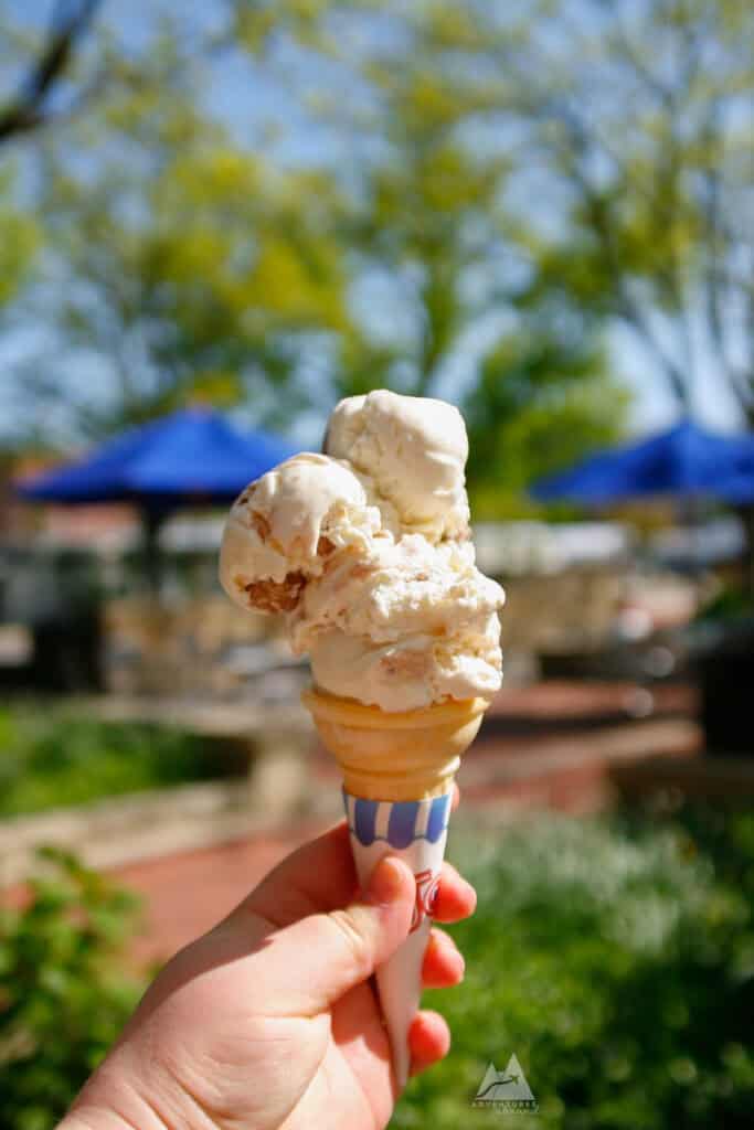 Getting ice cream from a local creamery like Berkey Creamery at Penn State is one of the top things to do in Happy Valley. A hand holds a waffle cone with scoops of creamy ice cream under bright sunlight. The ice cream appears to be a mix of vanilla with caramel swirls. Blue patio umbrellas and blurred greenery are visible in the background, suggesting an outdoor setting.