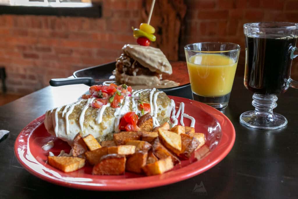 A hearty brunch plate featuring a smothered burrito topped with green sauce, sour cream, and fresh pico de gallo, served with crispy home fries. In the background, a skillet dish with a biscuit and gravy, a glass of orange juice, and a cup of black coffee are visible. The meal is set against a rustic brick wall, creating a cozy atmosphere at Creekside at Gamble Mill in Bellefonte, PA.