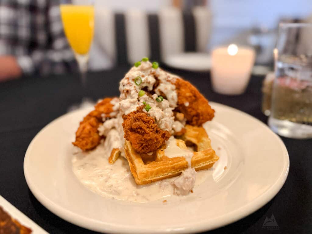 Brunch at GiGi’s Southern Table with a plate of chicken and waffles topped with creamy sausage gravy and garnished with chopped green onions. The golden, crispy fried chicken sits atop fluffy waffles, with a mimosa in the background. A softly lit candle and a blurred dining setting add a warm, inviting touch.