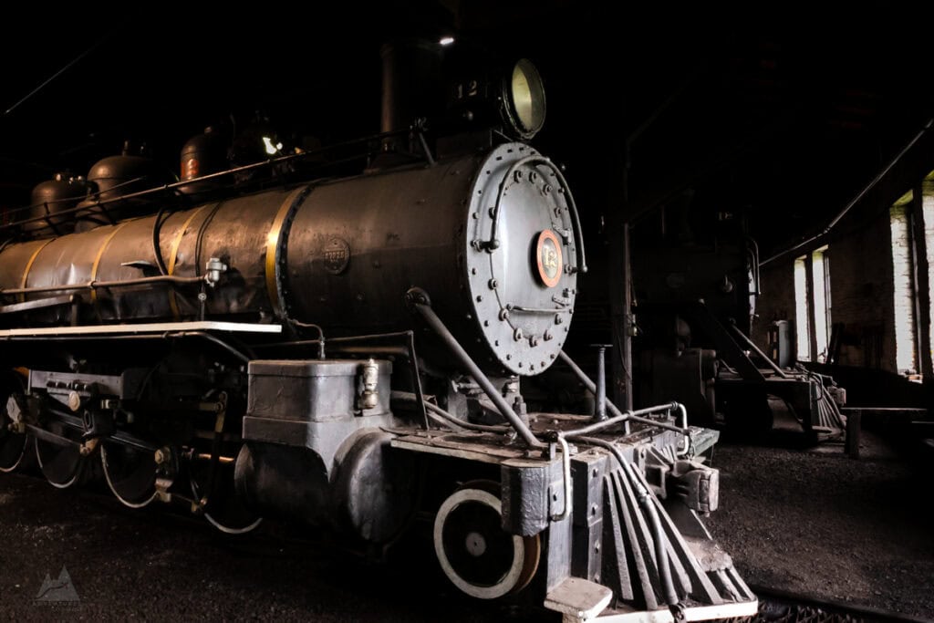 A vintage black steam locomotive rests inside a dimly lit rail yard at the historic East Broad Top Railroad. The large, round front plate, metal rivets, and aged details showcase its historical significance, with faint light streaming in through small windows in the background.
