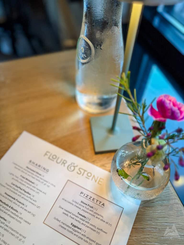 A cozy dining scene featuring a menu from "Flour & Stone" on a wooden table, accompanied by a glass bottle of chilled water and a small vase with pink flowers. Soft lighting and a window in the background add to the ambiance of this fine dining farm-to-table restaurant.