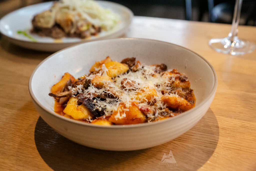 A close-up of a bowl of freshly made gnocchi, topped with shredded cheese, mushrooms, and short-rib. The dish sits on a wooden table, with another plate of food slightly out of focus in the background.