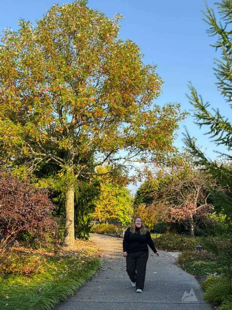 A woman in black clothing walks along a paved garden path at the H.O. Smith Botanic Gardens in State College, surrounded by colorful autumn foliage. A tall tree with mixed green and orange leaves towers over her, while neatly landscaped plants and shrubs frame the walkway.