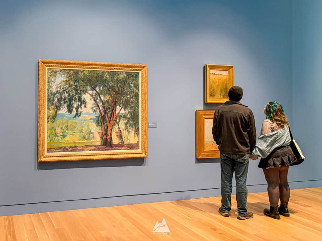 Palmer Museum at Penn State:
A couple stands in an art gallery, admiring a large impressionist painting of a tree and countryside, framed in gold. They are surrounded by additional framed artwork on a blue-gray wall. The gallery’s wood flooring and soft lighting create a calm, contemplative atmosphere.