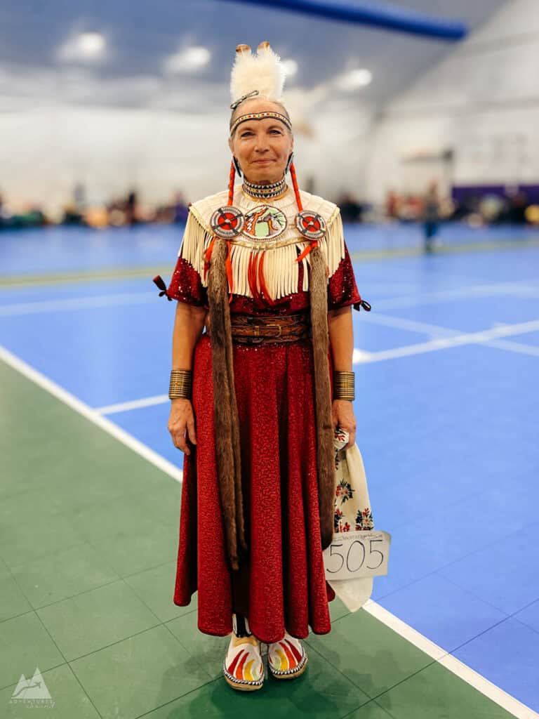 An older fair-skinned woman with long gray hair braided in pigtails is dressed in traditional Native American regalia stands on a gymnasium floor, wearing a red dress adorned with intricate beadwork, fringe, and fur details. She has a feathered headpiece, beaded jewelry, and moccasins with colorful embroidery. She holds a numbered card with "505" on it, indicating participation in a powwow event.