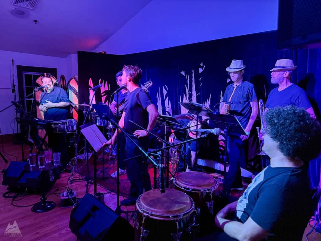 A Latin music band performs live on a dimly lit stage with a deep blue and purple glow. The band consists of musicians playing various instruments, including a keyboard, drums, saxophone, and trumpet. Two musicians wearing a fedora look toward the audience while others focus on the leader smiling and laughing towards them during a performance.