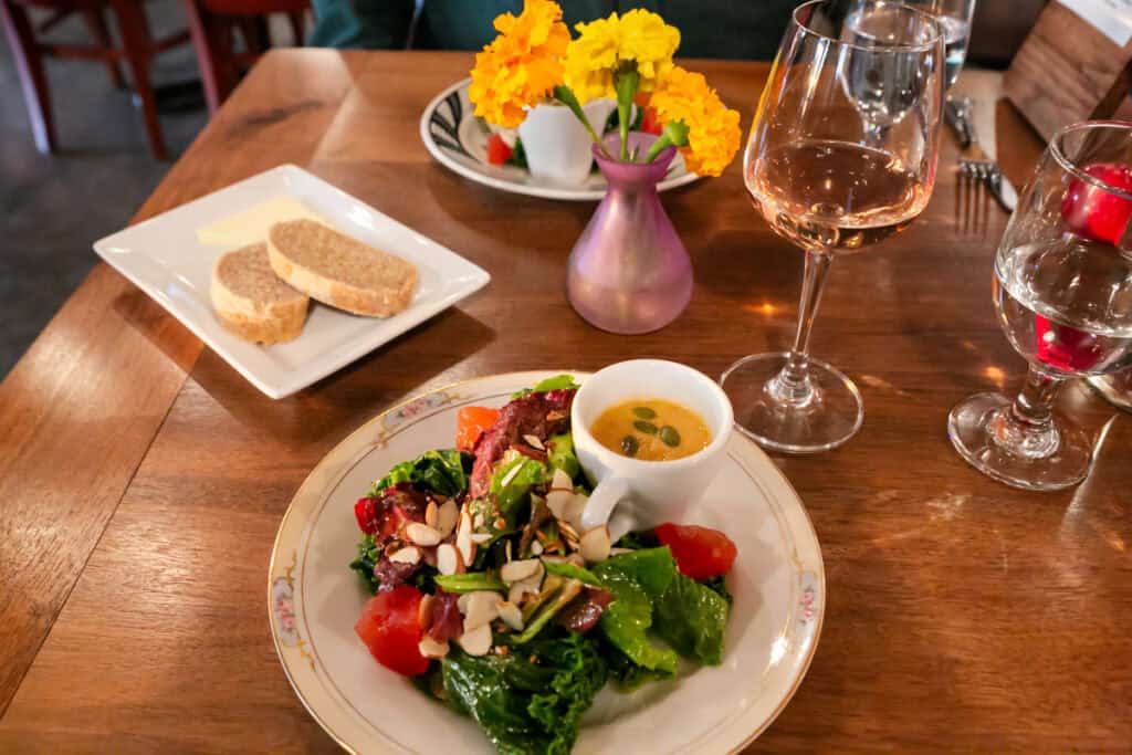 A beautifully plated farm-to-table meal sits on a wooden table, featuring a fresh salad with leafy greens, tomatoes, almonds, and a small cup of dressing. A plate of bread and butter is nearby, along with a glass of rosé wine and a vase of bright yellow marigold flowers. The cozy, warmly lit dining setting adds to the inviting atmosphere.