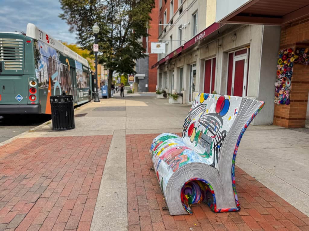 A uniquely designed public bench in the shape of an open book sits on a brick sidewalk in State College. The bench is painted with vibrant designs, including music notes and colorful shapes. A city bus with an advertisement is parked in the background, along with a row of buildings and a decorated storefront.