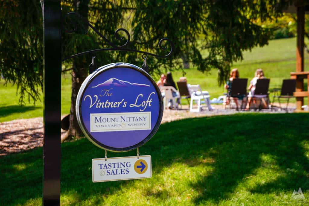 A decorative sign reading "The Vintner's Loft - Mount Nittany Vineyard & Winery" hangs from a metal bracket, with an additional sign below pointing towards "Tasting & Sales." In the background, a group of people relaxes on Adirondack chairs in a lush green outdoor setting, shaded by trees.