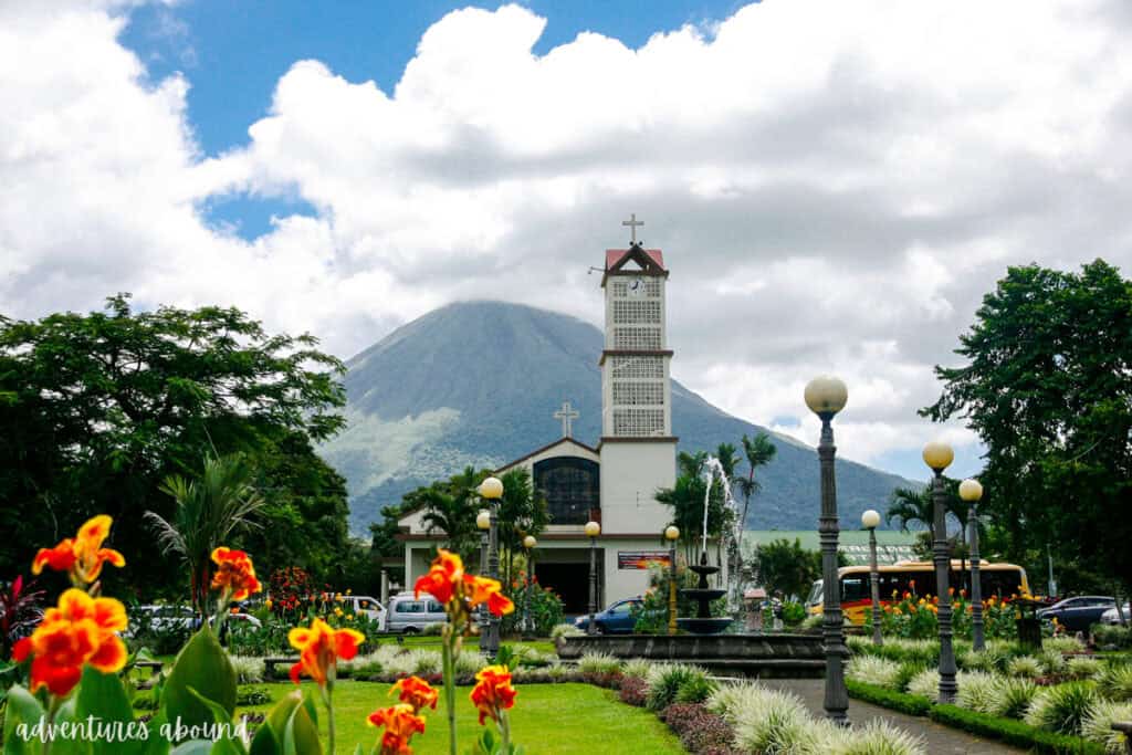 Arenal Volcano in Costa Rica rising into the clouds, with a customary Central American church in front of a square with red and yellow flowers and a fountain, all framing the volcano in the background.