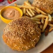 Rice Paper Cheeseburger Wellington with sesame seed bun, fries, and dipping sauce on a white plate.
