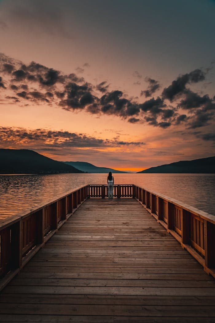 A tranquil sunset view from a wooden pier overlooking a calm lake and distant mountains.